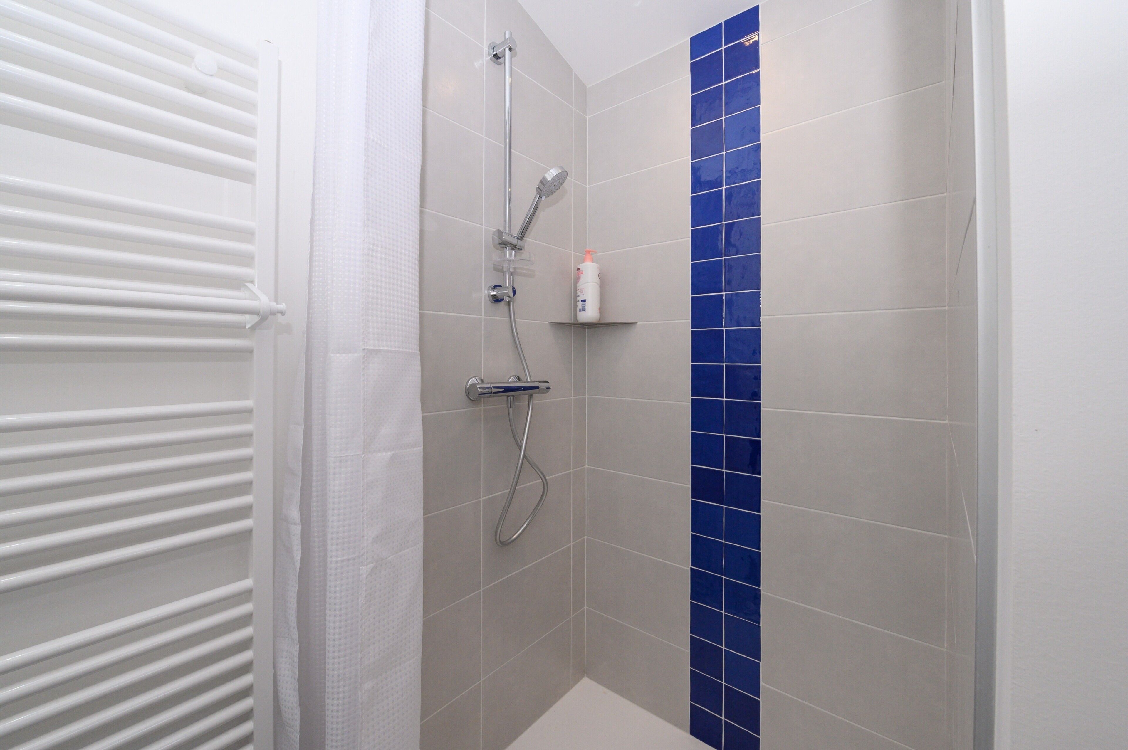 Eine Dusche mit Duschbrause und Wandarmatur, ein weißer Handtuchhalter und eine kleine Ablage mit einer Flasche.