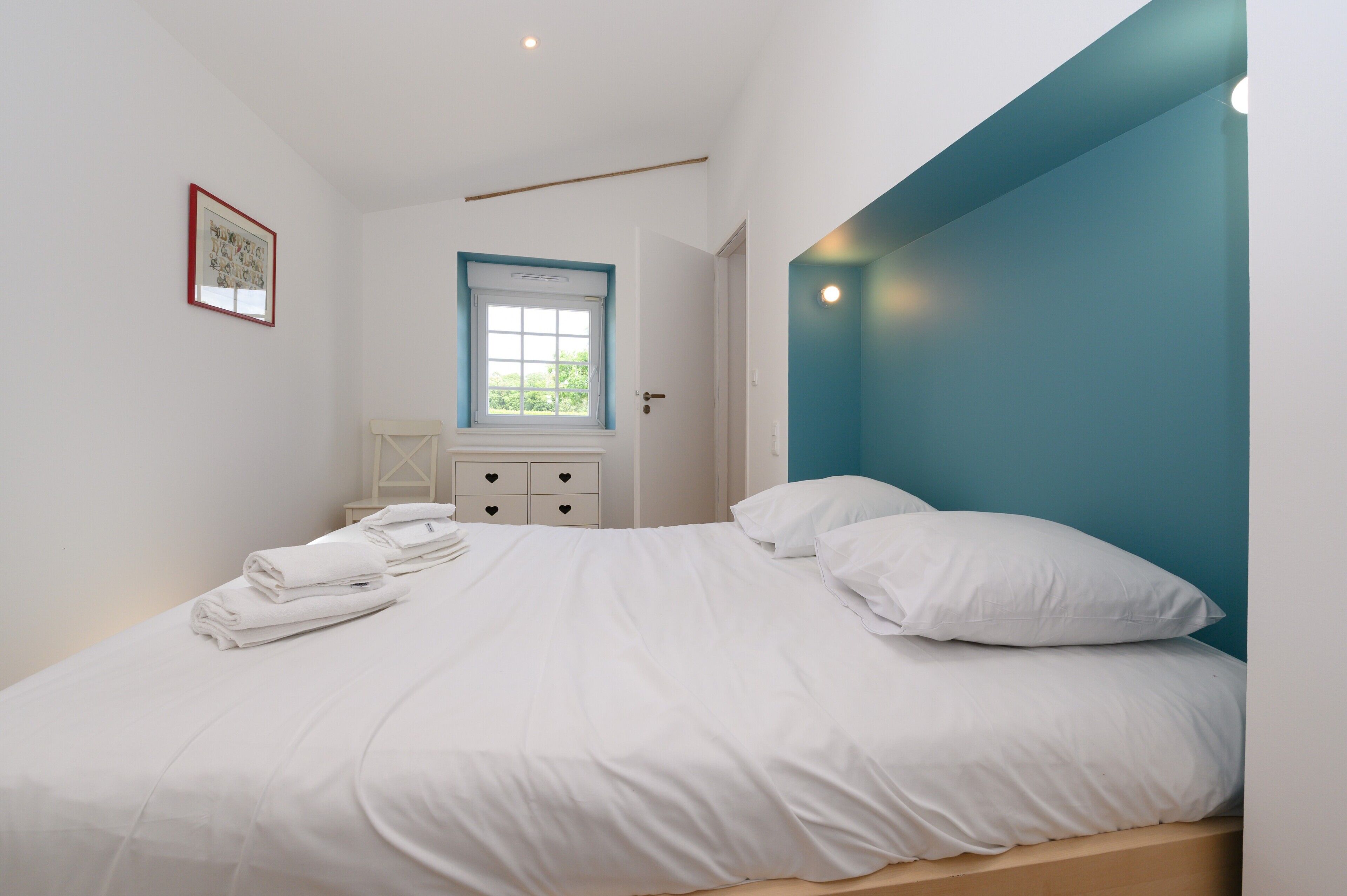 A bedroom with a bed, a window, a bedside table, and a wall-mounted picture.