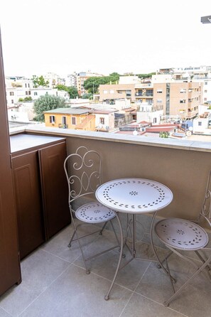 Ein Balkon mit zwei Metallstühlen und einem kleinen runden Tisch, mit Blick auf ein Wohngebiet mit Gebäuden und Bäumen.