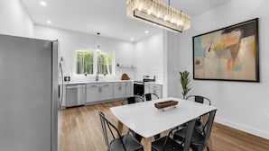 A modern kitchen with white cabinetry, a dining table with chairs, a chandelier, and a large abstract painting on the wall.