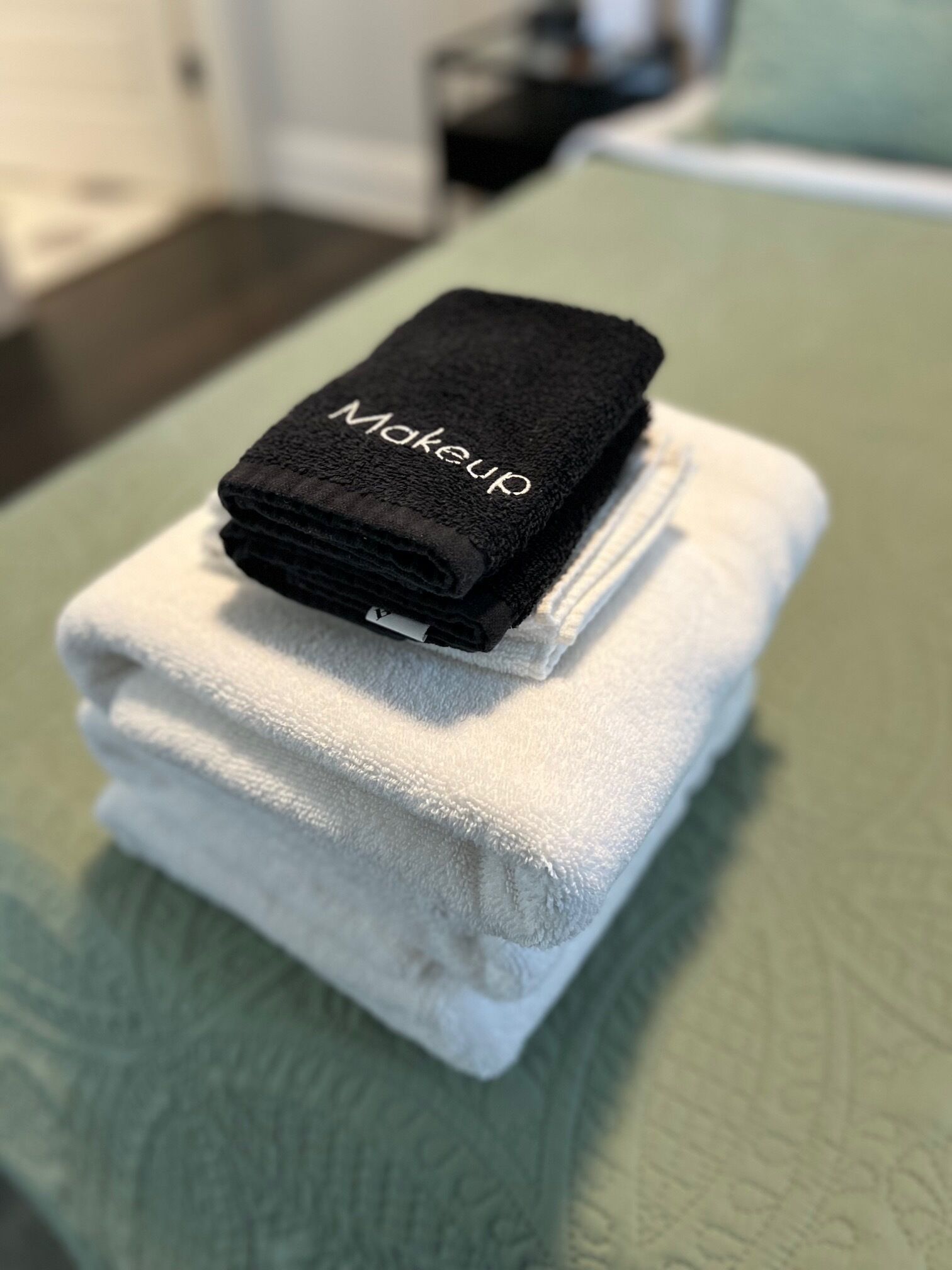 Una toalla negra doblada con la palabra 'Makeup' escrita, apilada sobre una toalla blanca en una cama con edredón verde.
