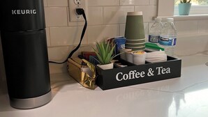 Un piano di lavoro in cucina con una macchina da caffè Keurig, un vassoio per caffè e tè, una pianta in vaso e una finestra vista alberi.