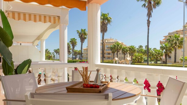 A sunlit outdoor seating area with a table, chairs, and a view of palm trees and buildings. - Marena Resort Townhouse (Oliva)