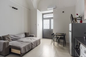 Ein kleines Zimmer mit einem Bett, einem Esstisch mit Stühlen, einer Küchenzeile mit Kühlschrank und einem Fenster mit Jalousien.