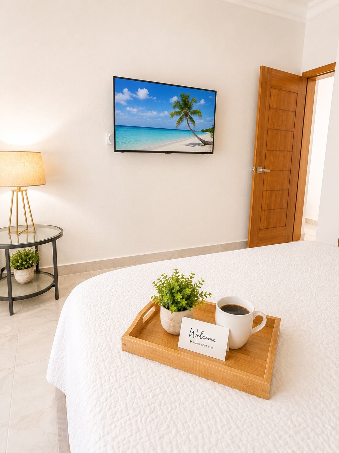 A hotel room with a bed, a welcome sign, a tray with a plant and a cup of coffee, a lamp, and a framed picture of a beach.