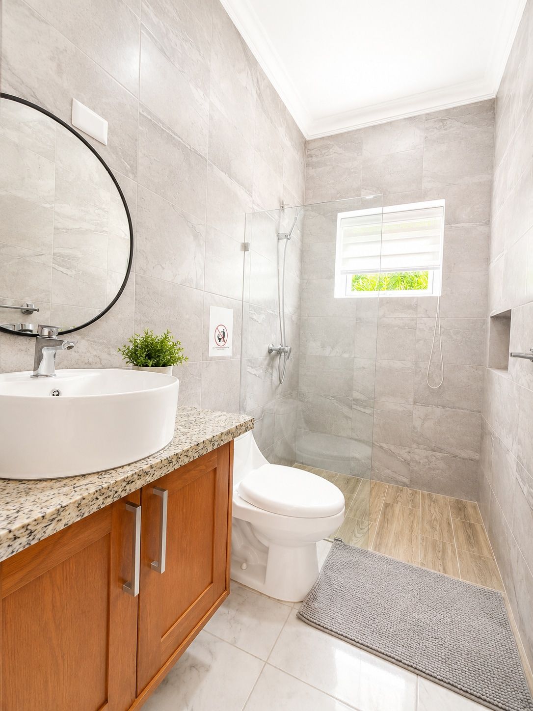 Un baño moderno con un lavabo redondo, un mueble de madera, inodoro, ducha y un pequeño ventanal con vistas.