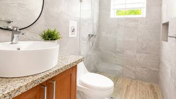 A modern bathroom with a round sink, wooden cabinet, toilet, shower, and a small window with a view.