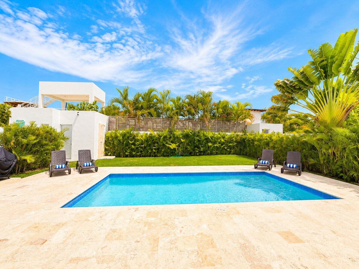 A swimming pool with lounge chairs, surrounded by greenery and a clear sky.