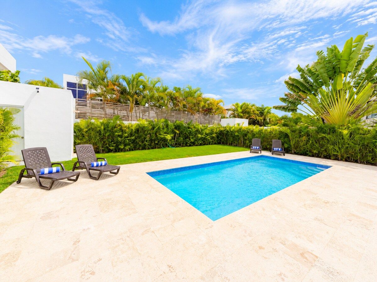 A rectangular swimming pool with a blue interior, surrounded by a light-colored patio. There are two lounge chairs with blue cushions and a small table. The pool is set against a backdrop of green foliage and a clear sky.