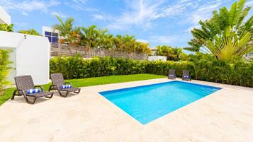 A rectangular swimming pool with a blue interior, surrounded by a light-colored patio. There are two lounge chairs with blue cushions and a small table. The pool is set against a backdrop of green foliage and a clear sky.