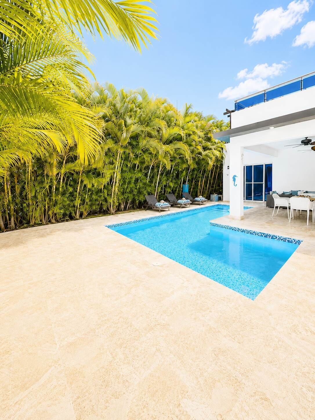 A swimming pool with a tiled deck, surrounded by palm trees and a modern building with a balcony.