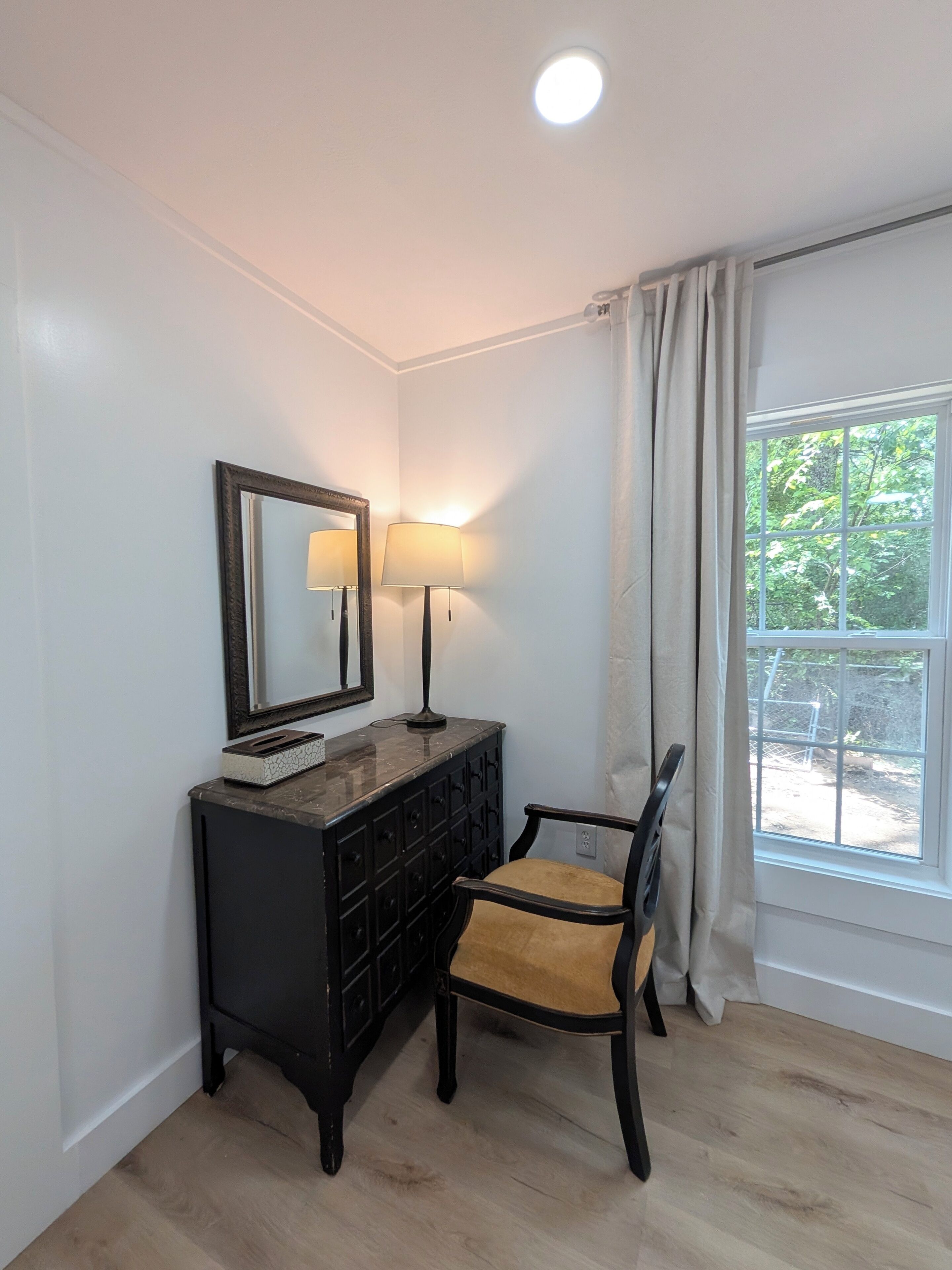 A room with a wooden floor, a black dresser with a mirror and lamps, a chair, and a window with curtains.
