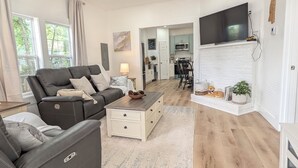 A modern living room with a ceiling fan, a sofa, a coffee table, a TV, and a kitchen area in the background. - Modern Historic Family Retreat House in Downtown Tupelo (Tupelo)