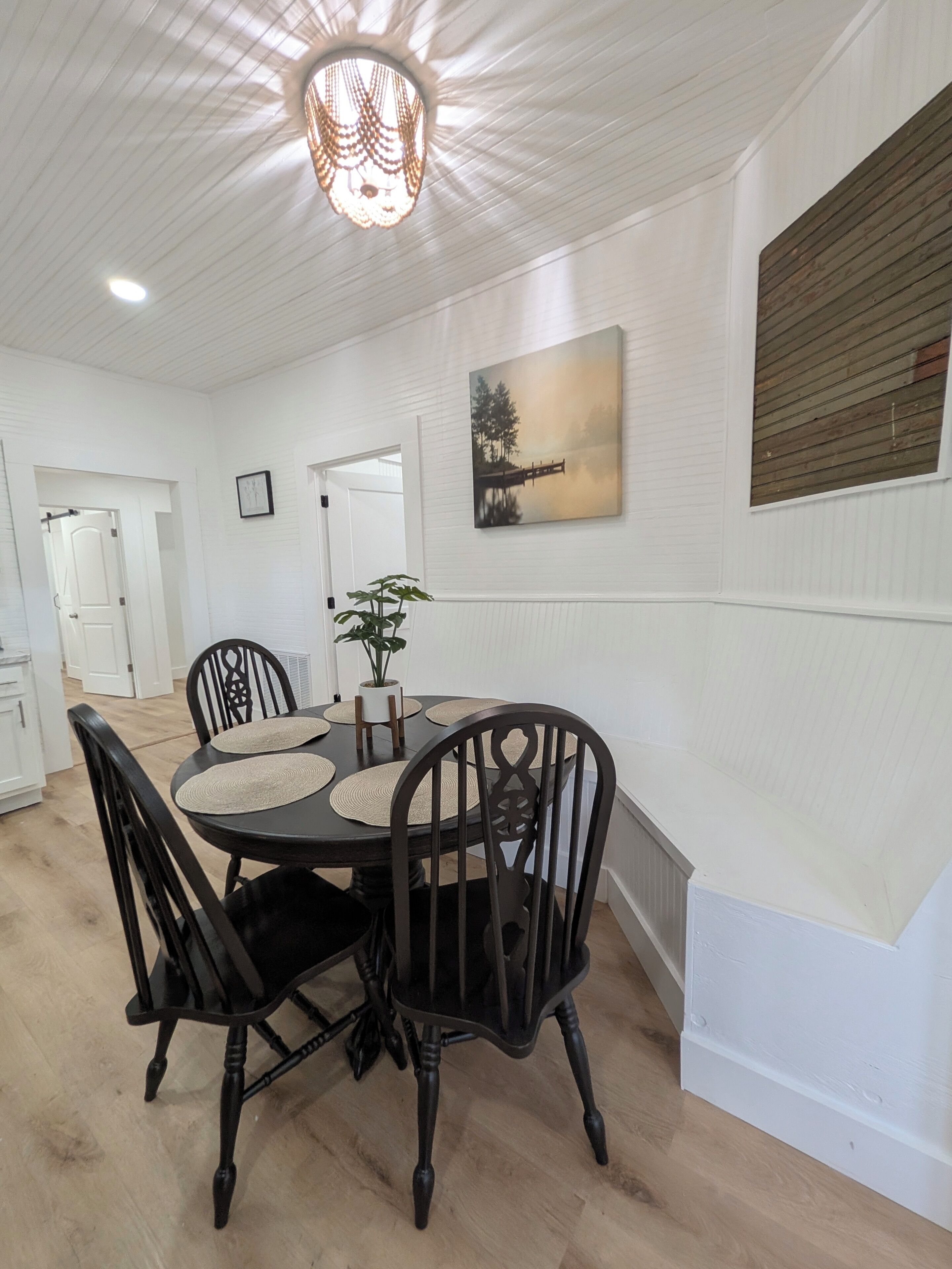 A dining area with a round table, black chairs, a potted plant, a painting, and a wooden panel on the wall.
