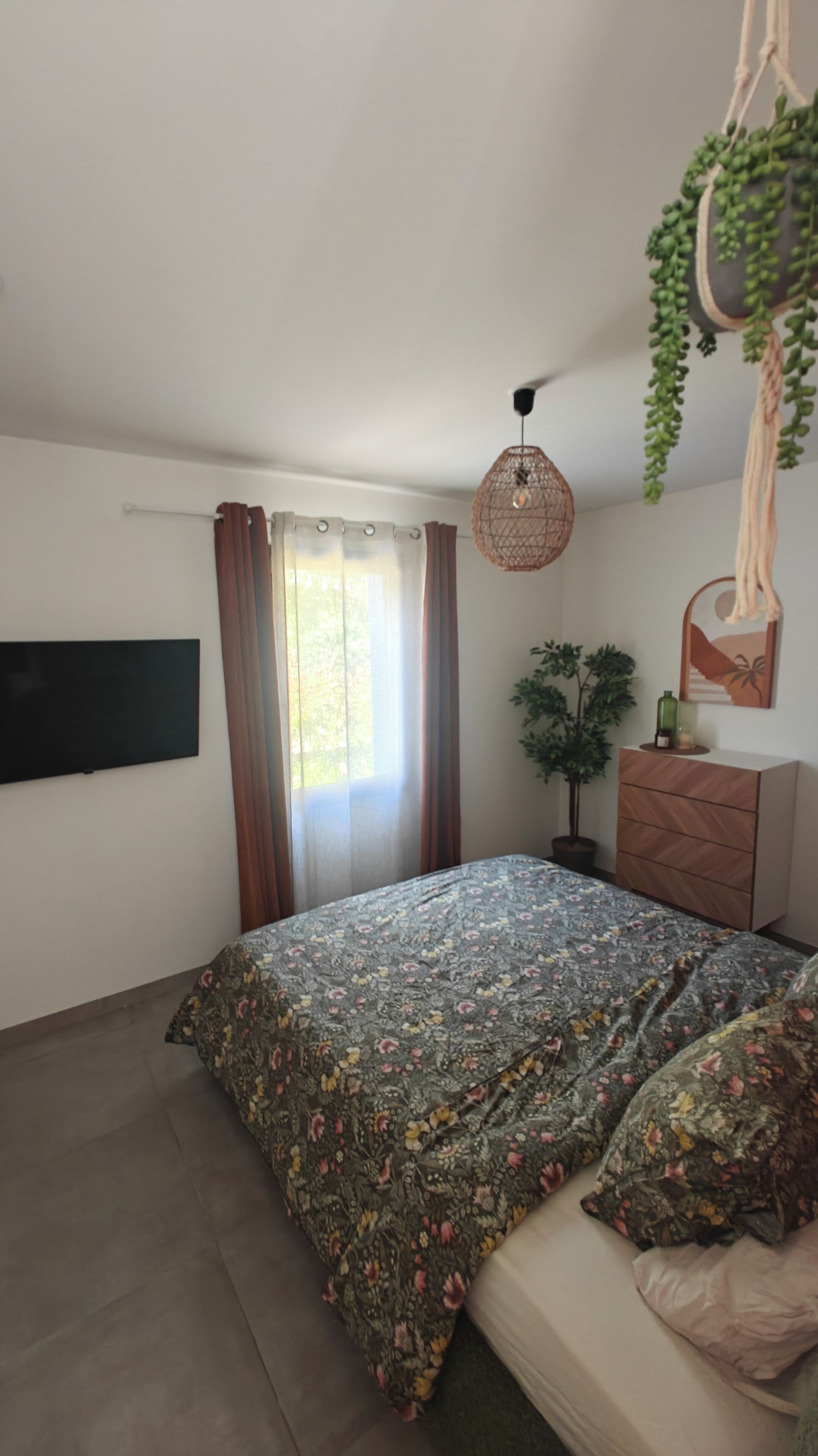 A bedroom with a bed, a TV, a hanging light fixture, a plant, and a dresser.