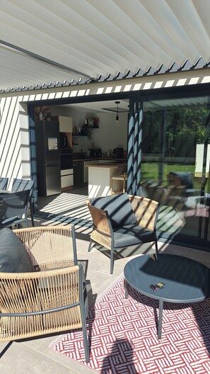 A modern outdoor seating area with wicker furniture and a patterned rug, a glass door leading to a kitchen, and a view of greenery. - CASA NUBA - Modern villa with Jacuzzi - Between Provence & Mont Ventoux (Carpentras)