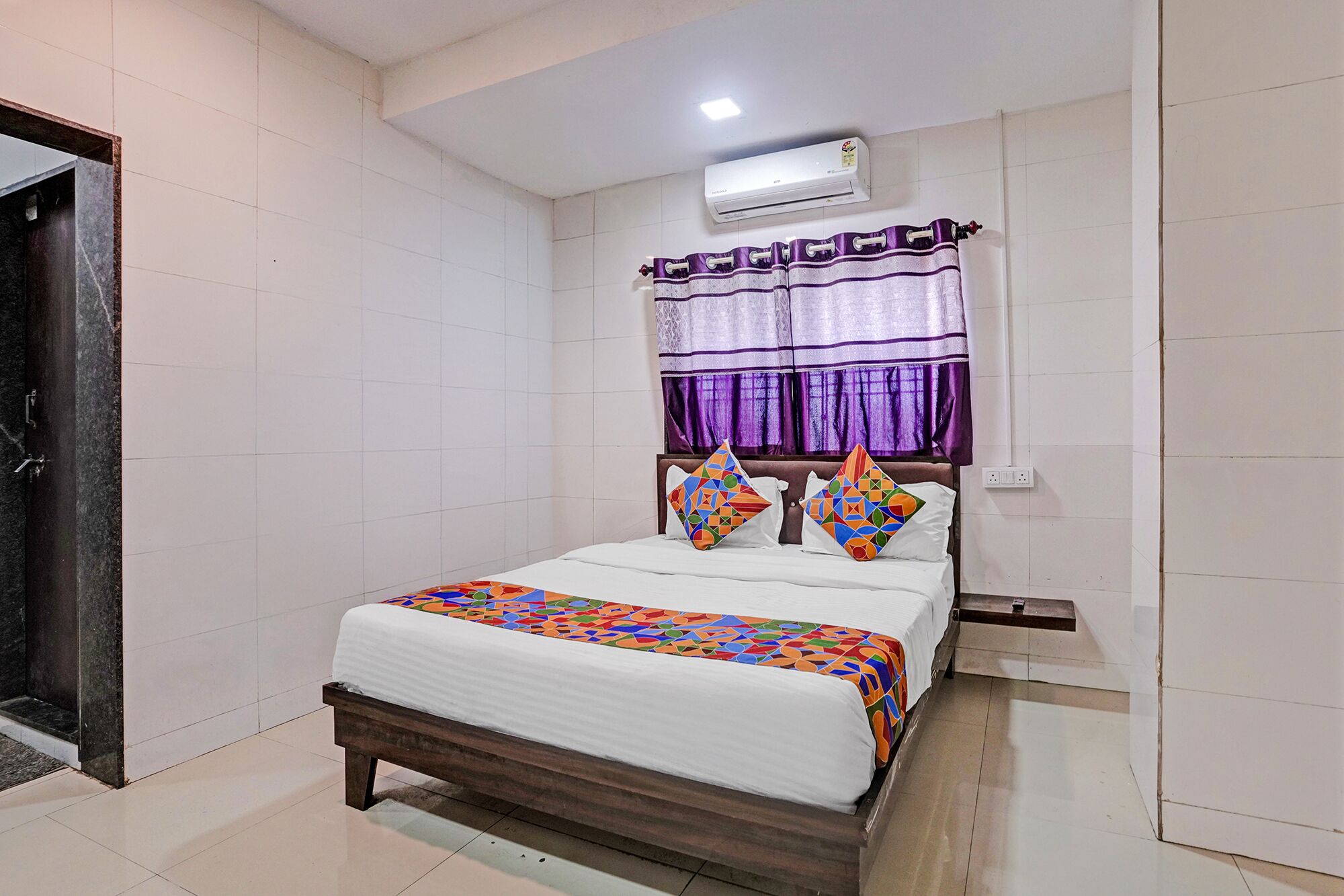 Un dormitorio con cama, ventana con cortinas moradas, aire acondicionado y mesita de noche.