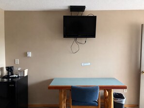 Ett rum med en tv som är monterad på väggen, ett bord med en stol, en kaffebryggare och en papperskorg.