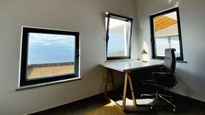 Una stanza con una scrivania, due finestre vista mare, una sedia e una lampada.
