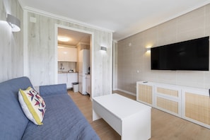 Ein modernes Wohnzimmer mit einer Couch, einem Couchtisch, einem flachbildfernseher an der Wand und einer durchgehenden Tür erkennbaren Küche.