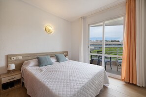 Ein Hotelzimmer mit Bett, Nachttisch, Lampe und Blick auf einen Balkon mit Sitzmöbeln.
