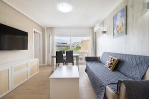 Ein modernes Wohnzimmer mit Flachbildfernseher, einer Couch mit gemustertem Überwurf, einem Essbereich mit Stühlen und Blick durch eine Glastür.