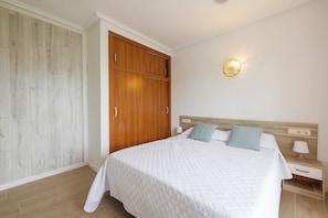 Ein Schlafzimmer mit einem Holzschrank, einem Bett mit weißer Steppdecke, zwei Kissen, einem Nachttisch mit Lampe sowie einer Wandleuchte.