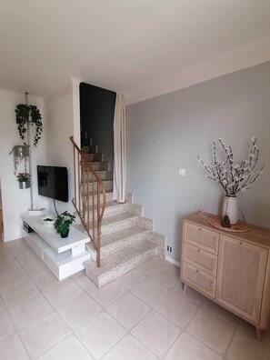 Una sala de estar con una escalera, un televisor de pantalla plana, un mueble blanco para equipo audiovisual, un armario de madera y un florero con ramas.