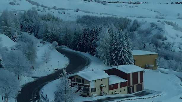 Paisaje nevado con un camino sinuoso, un pequeño edificio de fachada roja y beige, y una zona boscosa.