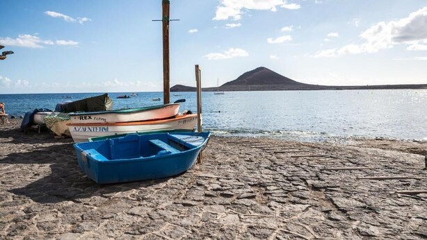 A beach with boats, a rocky shore, and a mountain in the background. - Las Olas (El Médano)