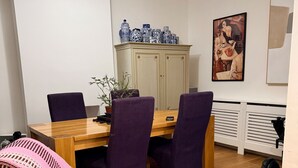 A dining area with a wooden table, purple chairs, a grey sofa with a pink and white blanket, a blue vase collection, a framed picture on the wall, and a radiator. - House Sleeps 2 With Parking (London)
