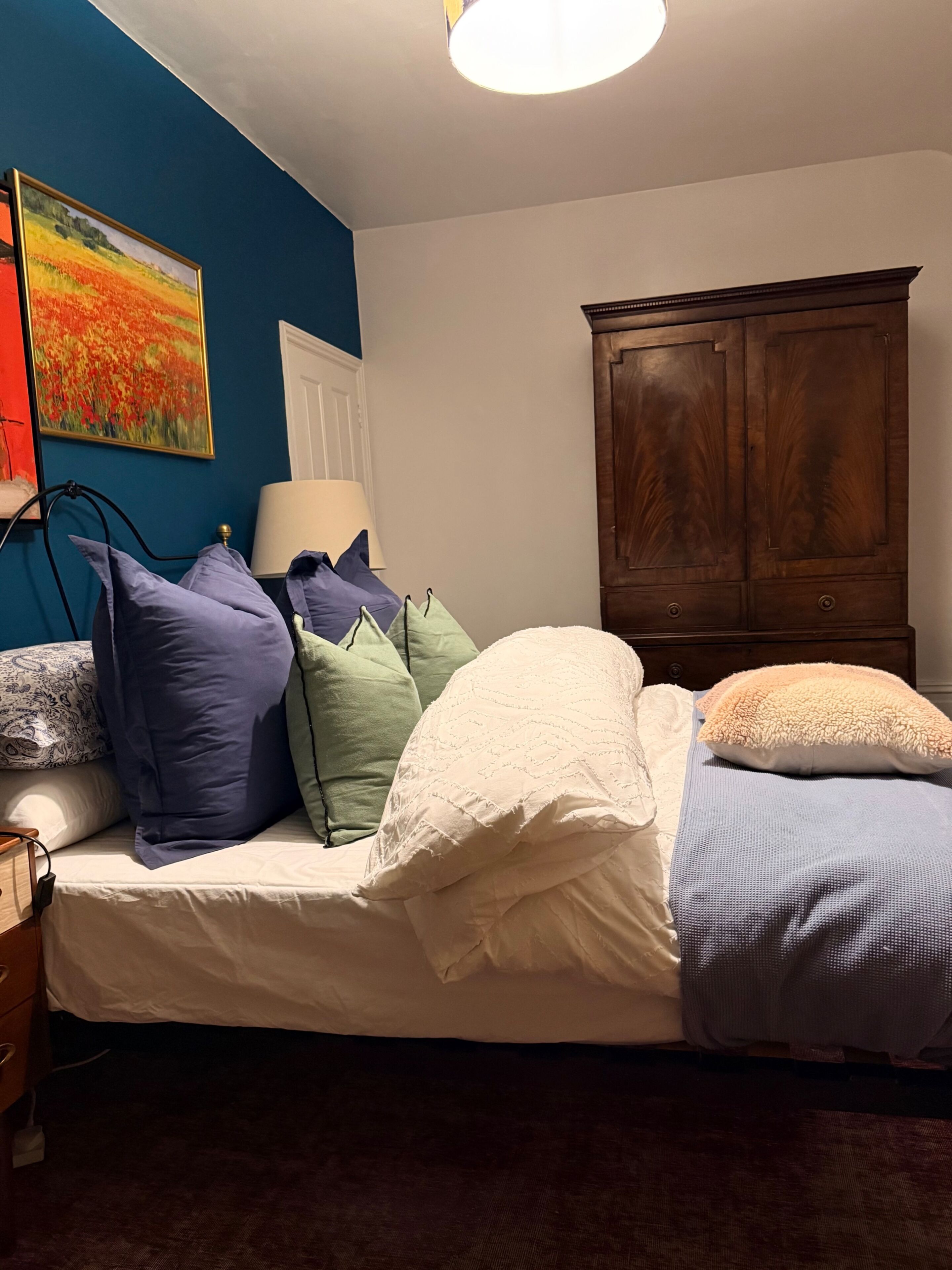 A bedroom with a bed, pillows, a lamp, a wooden cabinet, and a painting on the wall.