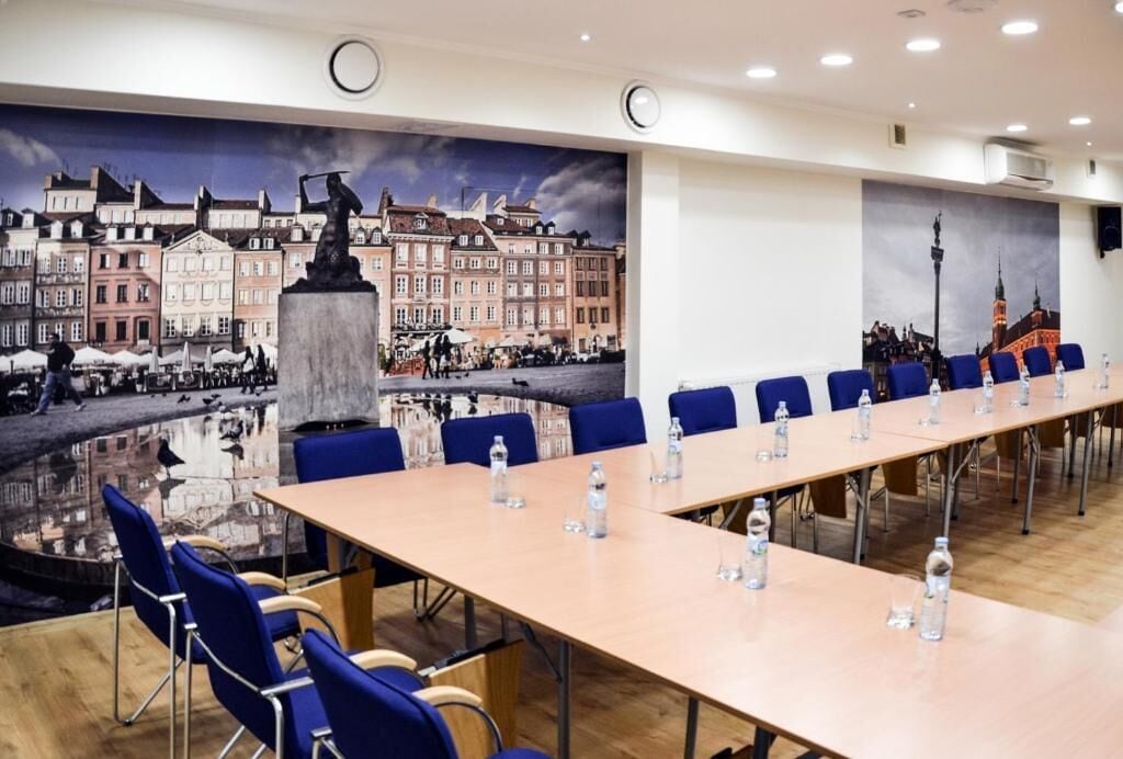 Ein Konferenzraum mit einem langen Tisch, blauen Stühlen und einem Wandbild einer europäischen Stadtlandschaft.