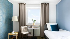 Una habitación de hotel con una cama, una silla, una mesita con una lámpara, una ventana con cortinas y una planta en el alféizar.