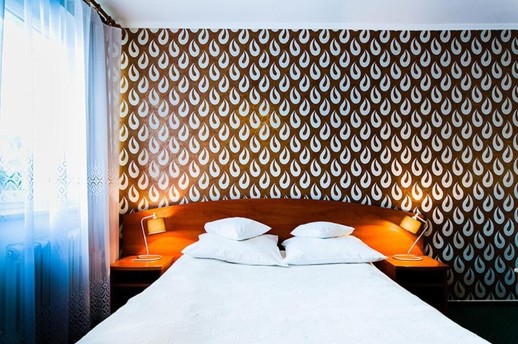 A hotel room with a patterned wallpaper, a bed with white linens, two bedside tables with lamps, and a window with curtains.