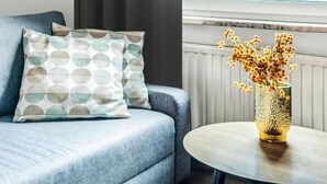 Una habitación con sofá gris, almohadones estampados, una mesa redonda con un jarrón texturizado y vistas al cielo a través de una ventana.