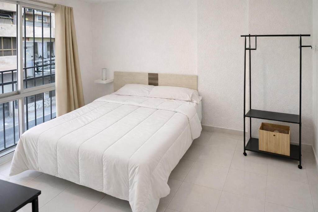 A hotel room with a bed, a window with curtains, a small table, and a black metal shelf with a wooden box.