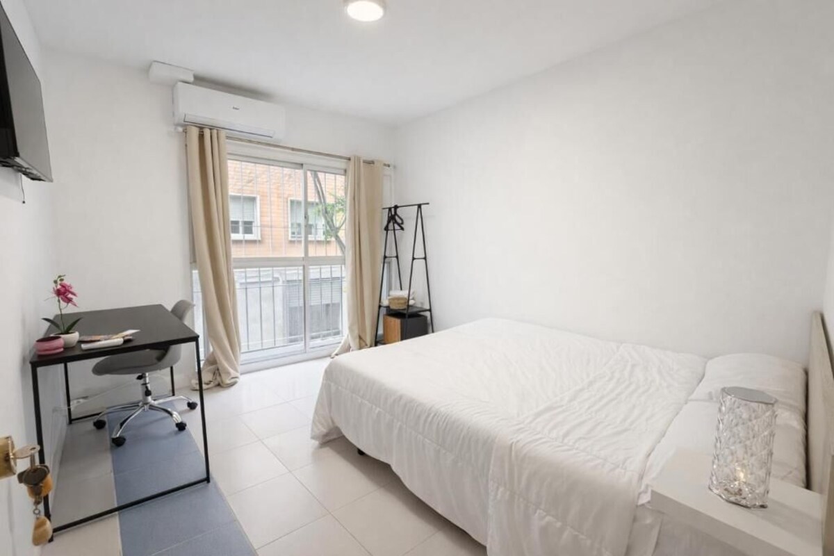 Un dormitorio con cama, escritorio, silla, lámpara colgante y ventana con cortinas.