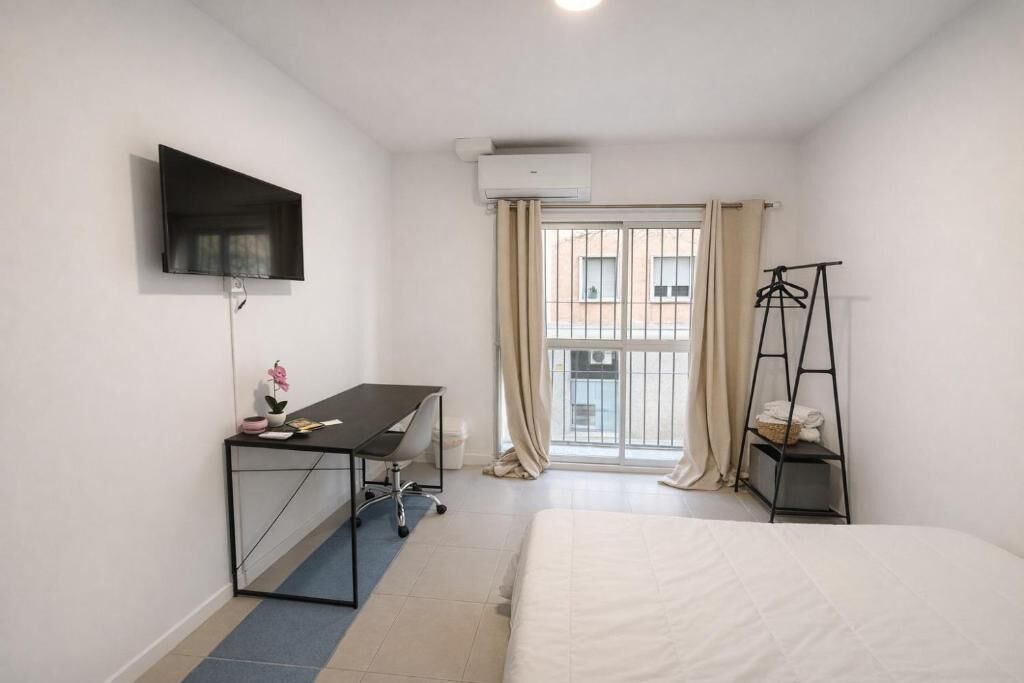 Habitación de hotel con cama, escritorio con silla, televisor montado en la pared, equipo de aire acondicionado, ventana con cortinas y un tendedero.