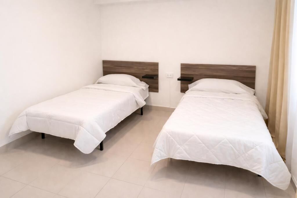 Dos camas individuales con cabeceros de madera en una habitación con paredes blancas y suelo de baldosas.