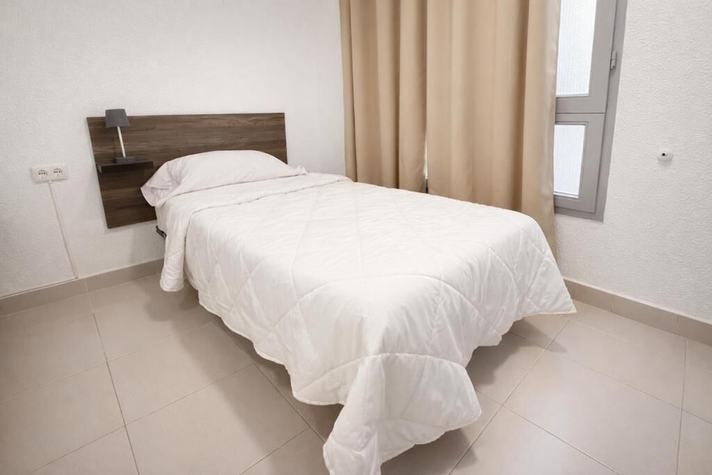 A single bed with a wooden headboard, beige curtains, and a window with a white frame.