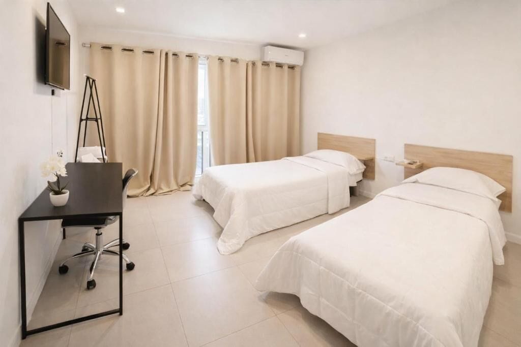 A hotel room with two beds, a desk with a chair, a TV mounted on the wall, and a window with curtains.