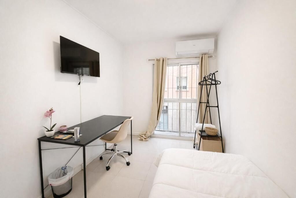Une petite pièce propre, comprenant un lit, un bureau, une chaise, un téléviseur, une fenêtre avec des rideaux et un étendoir à linge.