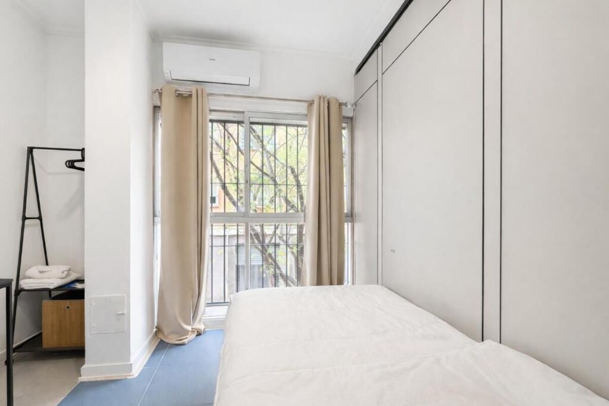 Un dormitorio con cama, ventana con cortinas, unidad de aire acondicionado y una mesita con una toalla.