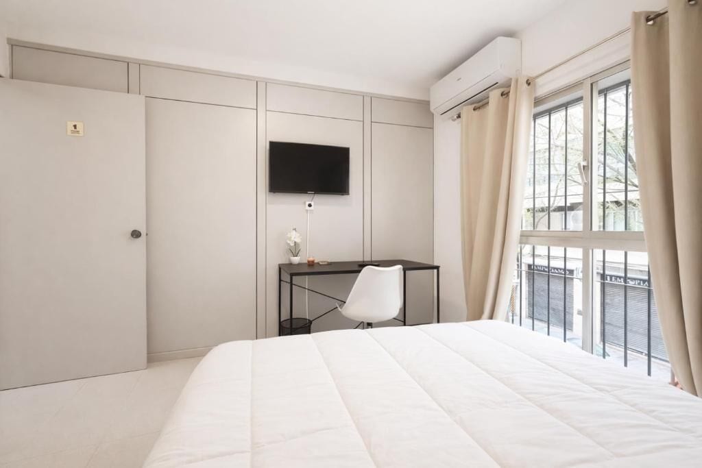 A bedroom with a bed, a desk, a chair, a TV, and a window with curtains.