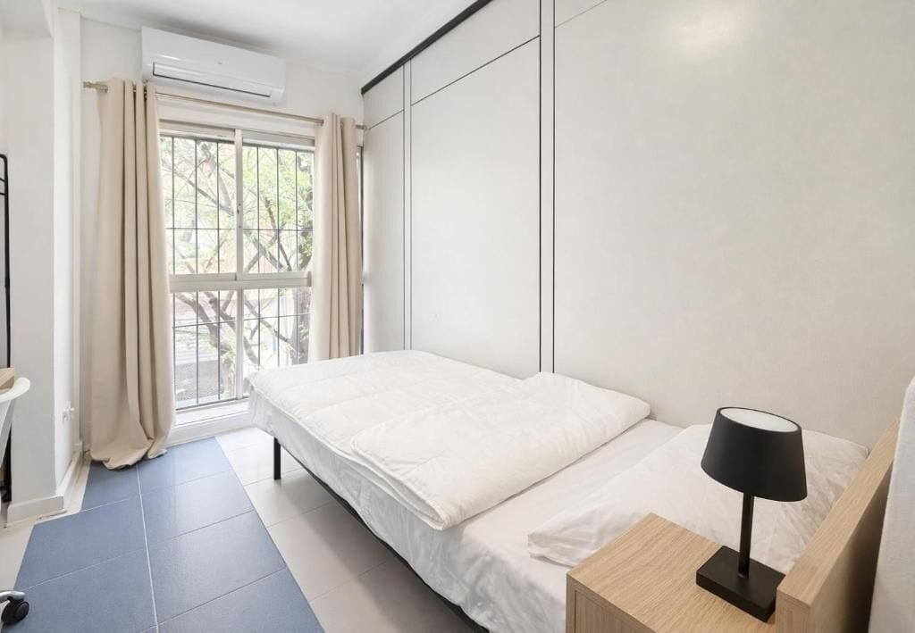 Quarto com cama, mesa de cabeceira com luminária, janela com cortinas e aparelho de ar condicionado.