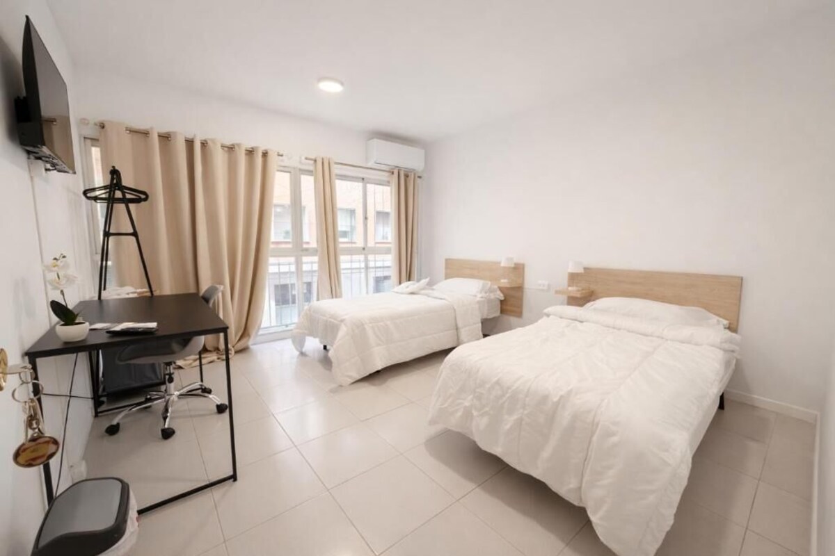 Habitación de hotel con dos camas, un escritorio con silla, un televisor montado en la pared y una ventana con cortinas.