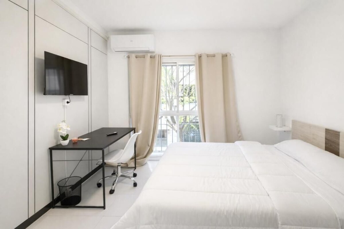 Una habitación de hotel con cama, escritorio con silla, televisor montado en la pared y ventana con cortinas.