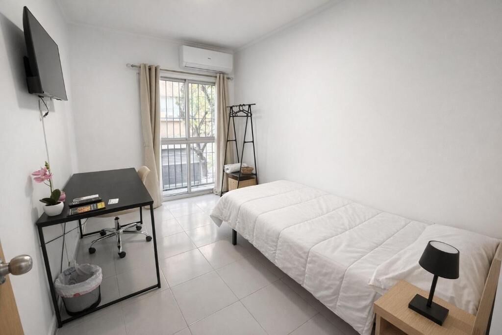Habitación de hotel con cama, escritorio con silla, televisor montado en la pared, ventana con cortinas y un equipo de aire acondicionado.