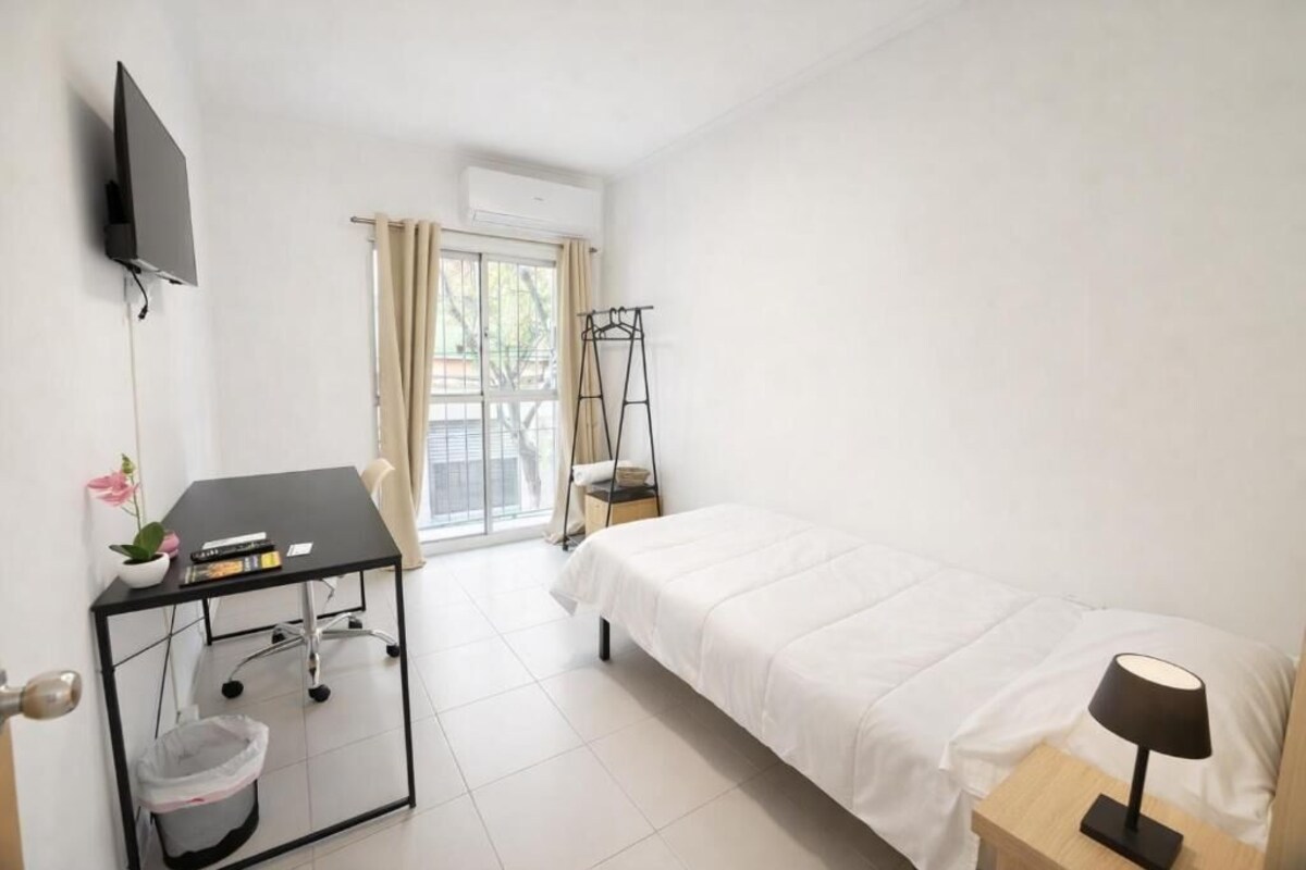 Habitación de hotel con cama, escritorio con silla, televisor, ventana con cortinas y una mesita con lámpara.